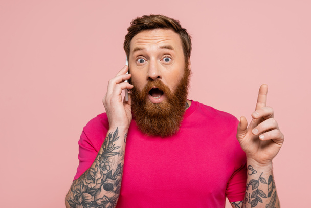 asombrado hombre tatuado hablando en el teléfono móvil y mostrando idea signo aislado en rosa - Foto, Imagen