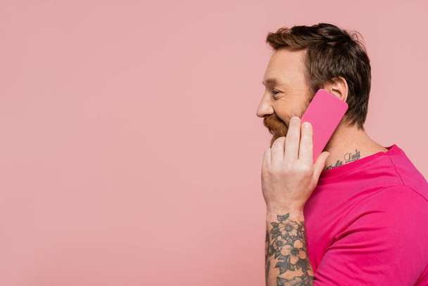 Вид сбоку жизнерадостного мужчины в майке маджента, звонящего по мобильному телефону, изолированному на розовом - Фото, изображение