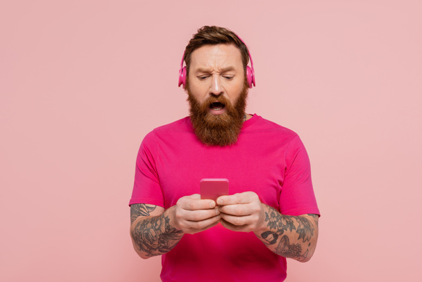 Kablosuz kulaklık takan şok olmuş sakallı adam pembe ile izole edilmiş cep telefonu kullanıyor. - Fotoğraf, Görsel