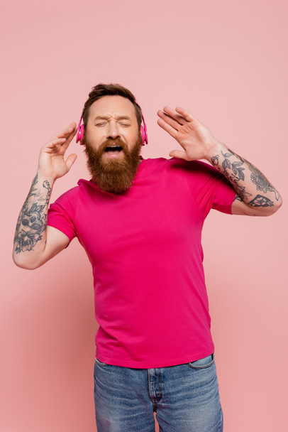 возбужденный бородатый человек слушает музыку в беспроводных наушниках и поет с закрытыми глазами изолированы на розовый - Фото, изображение