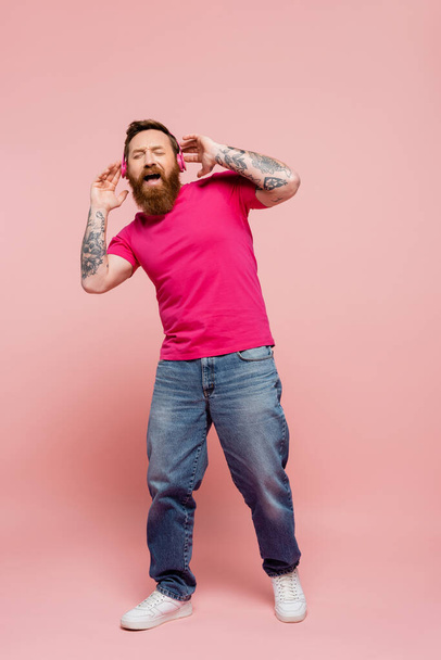longitud completa del hombre asombrado con los ojos cerrados escuchando música en auriculares inalámbricos y cantando sobre fondo rosa - Foto, imagen