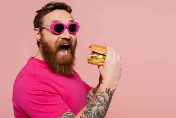 thrilled man in trendy sunglasses opening mouth near tasty burger isolated on pink - Valokuva, kuva