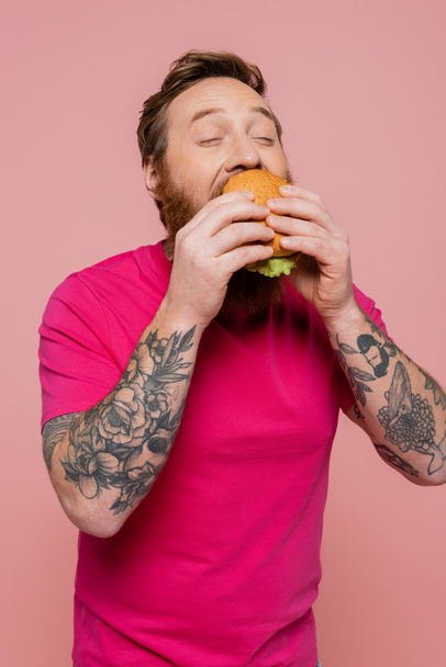 hombre tatuado complacido con los ojos cerrados comiendo deliciosa hamburguesa aislada en rosa - Foto, Imagen
