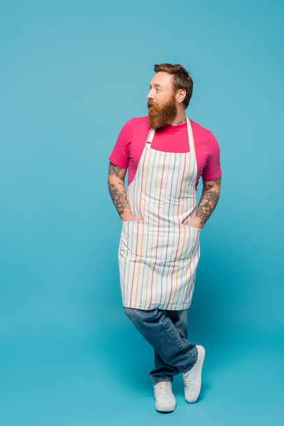 full length of tattooed man standing with hands in pockets of striped apron and looking away on blue background - Foto, imagen