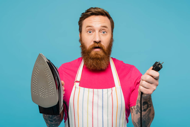 discouraged bearded man in striped apron holding iron and looking at camera isolated on blue - Foto, immagini
