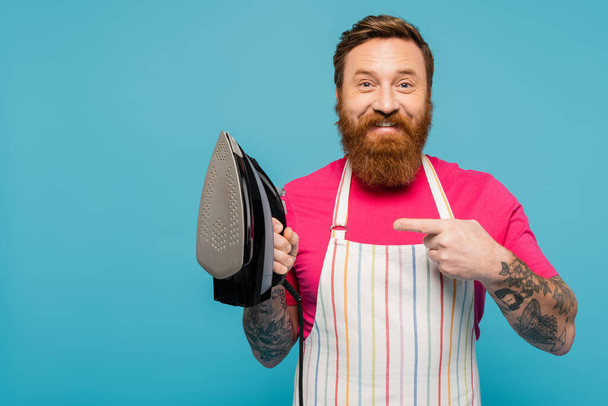 happy tattooed man in striped apron looking at camera while pointing at iron isolated on blue - Fotoğraf, Görsel