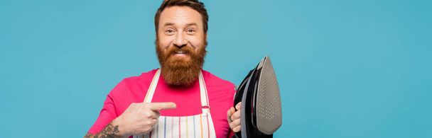 cheerful bearded man in apron pointing at iron and looking at camera isolated on blue, banner - Фото, зображення