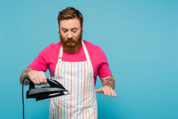 bearded man in magenta t-shirt and striped apron holding iron isolated on blue - Foto, afbeelding