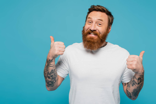 happy bearded and tattooed man in white t-shirt showing thumbs up and looking at camera isolated on blue - Fotografie, Obrázek