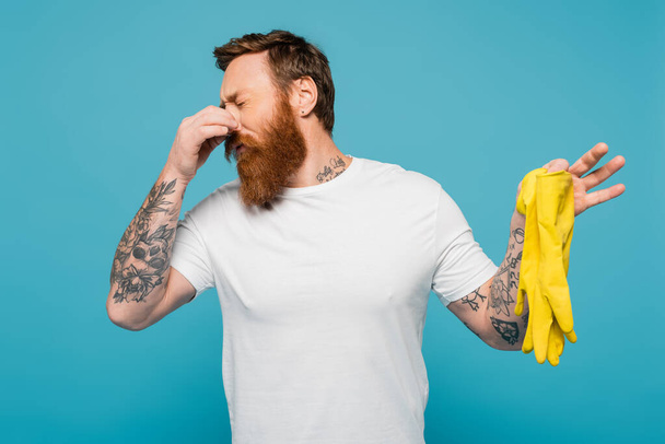 disgusted bearded man in white t-shirt plugging nose while holding stinky rubber gloves isolated on blue - Foto, Imagem