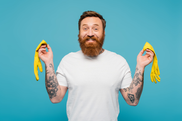 joyful tattooed man in white t-shirt looking at camera while holding yellow rubber gloves isolated on blue - Foto, Imagem