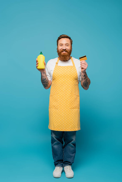 full length of happy bearded man in polka dot apron posing with credit card and detergent on blue background - Фото, изображение