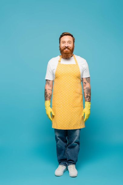 full length of bearded tattooed man in yellow apron and rubber gloves smiling at camera on blue background - Fotó, kép