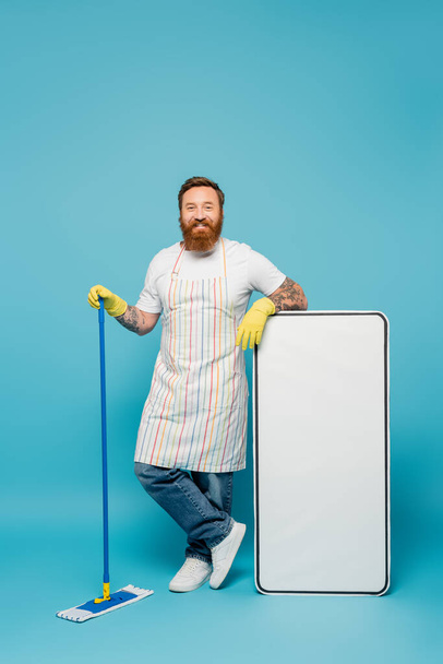 cheerful tattooed man in yellow rubber gloves and striped apron holding mop near huge smartphone template on blue background - Фото, зображення