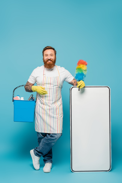 homem barbudo alegre em avental listrado de pé com escova de poeira e balde perto modelo de telefone branco no fundo azul - Foto, Imagem