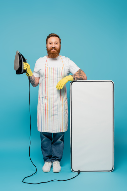 hombre barbudo alegre en delantal y guantes de goma posando con hierro cerca de la maqueta del teléfono grande sobre fondo azul - Foto, imagen