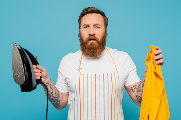 tense bearded man in striped apron holding yellow cloth and iron isolated on blue - Fotoğraf, Görsel