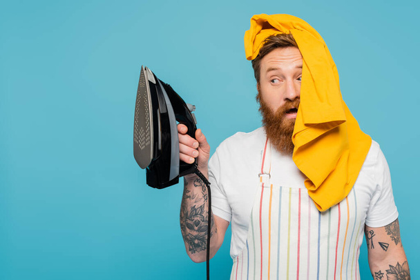 bearded man with yellow garment on head looking at electric iron isolated on blue - Photo, Image