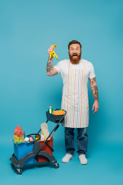 amazed bearded man in apron holding rubber gloves near cart with cleaning supplies on blue background - Photo, Image