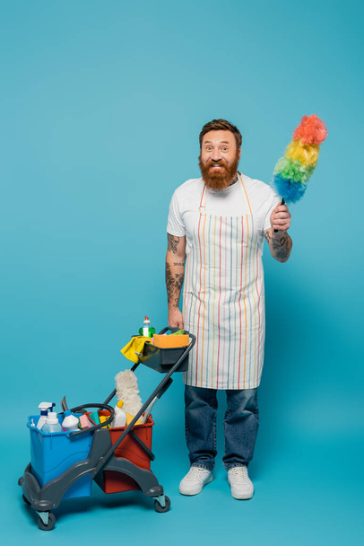 cheerful man in apron posing with dust brush near cart with cleaning supplies on blue background - Foto, Imagem