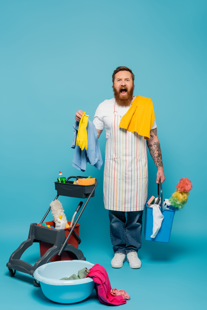 stressed bearded man in striped apron screaming near laundry and cleaning supplies on blue background - Foto, Imagem
