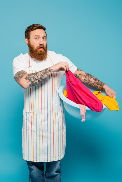 discouraged bearded man in apron holding laundry bowl and looking at camera isolated on blue - Valokuva, kuva