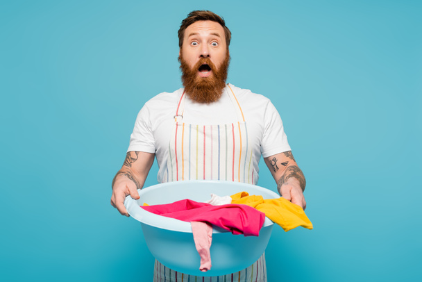shocked man in striped apron holding bowl with laundry and looking at camera isolated on blue - Foto, Imagem
