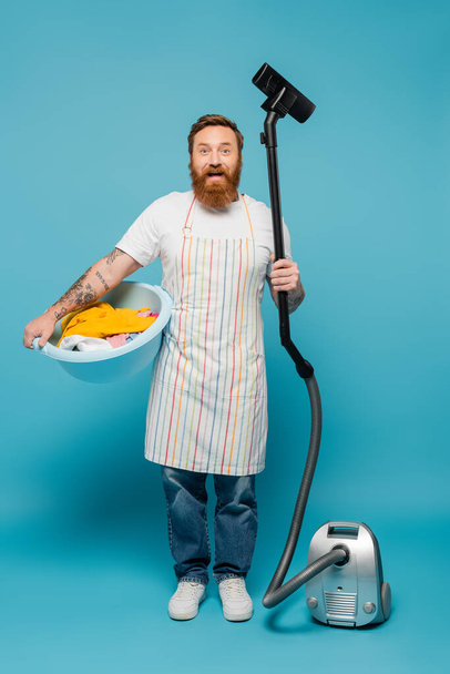 joyful tattooed man in apron holding laundry bowl and vacuum cleaner while looking at camera on blue background - Photo, image