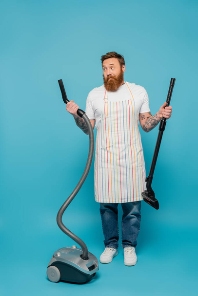 full length of discouraged man in striped apron looking at parts of vacuum cleaner on blue background - Фото, зображення