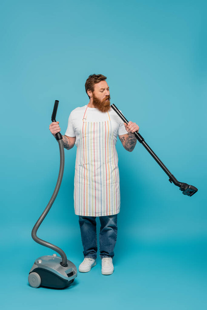 full length of thoughtful man in striped apron looking in tube of vacuum cleaner on blue background - Foto, Imagen