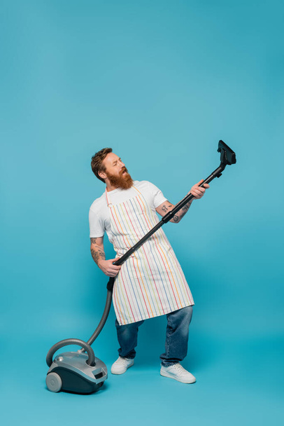 comprimento total de homem barbudo animado em avental listrado se divertindo com aspirador de pó no fundo azul - Foto, Imagem