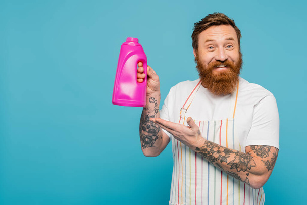 joyful bearded man in striped apron pointing at bottle with washing gel isolated on blue - Fotoğraf, Görsel