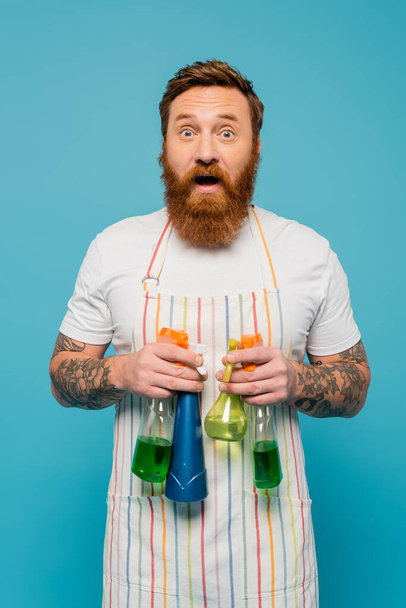 hombre barbudo emocionado en delantal rayado sosteniendo diferentes botellas de aerosol y mirando a la cámara aislada en azul - Foto, imagen