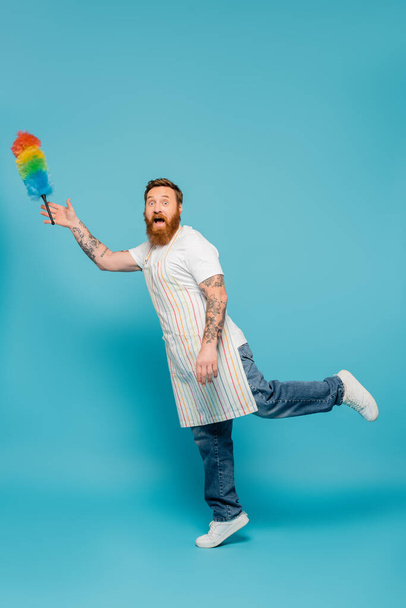 full length of amazed bearded man in striped apron looking at camera near levitating dust brush on blue background - Foto, Imagen