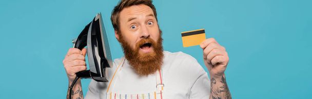 zdumiony brodaty mężczyzna pokazując żelaza i karty kredytowej izolowane na niebieski, sztandar - Zdjęcie, obraz