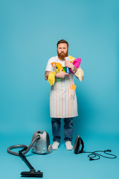 full length of serious tattooed man holding detergents near vacuum cleaner and iron on blue background - Photo, Image