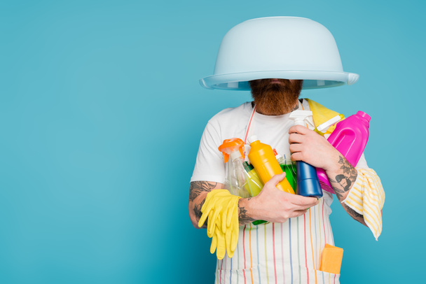 γενειοφόρος άνθρωπος με διαφορετικές προμήθειες καθαρισμού και μπολ πλυντηρίου στο κεφάλι απομονώνονται σε μπλε - Φωτογραφία, εικόνα