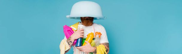 tattooed man with cleaning supplies and laundry bowl on head isolated on blue, banner - Foto, Bild
