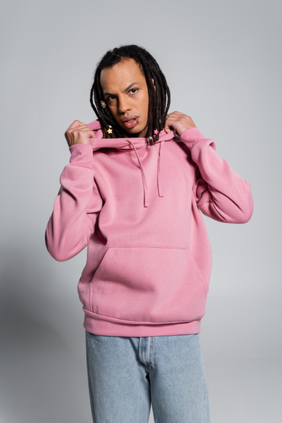 pierced multiracial man with dreadlocks adjusting pink hoodie on grey  - Foto, immagini
