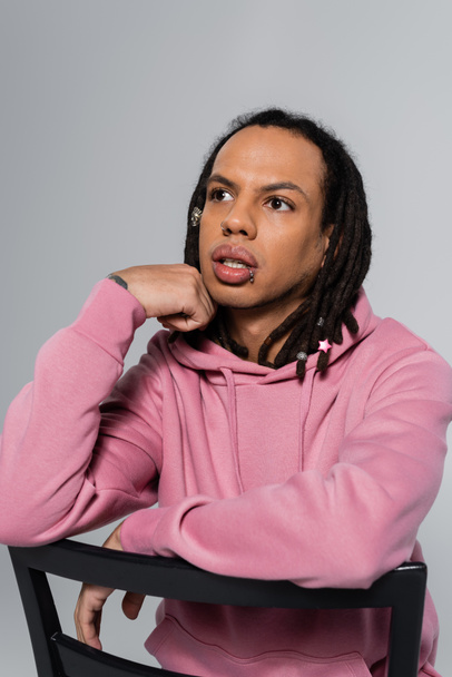 thoughtful multiracial man in pink hoodie leaning on chair back isolated on grey  - Valokuva, kuva