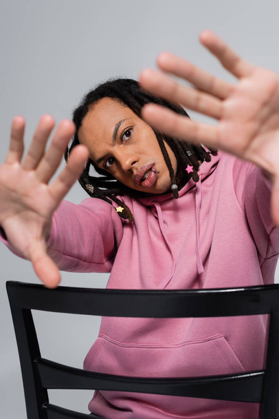 pierced multiracial man in pink hoodie gesturing while sitting on chair isolated on grey  - Photo, Image