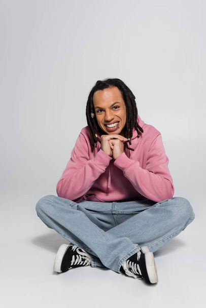 full length of happy multiracial man with dreadlocks sitting with crossed legs on grey  - Foto, afbeelding