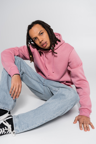full length of multiracial and pierced man with dreadlocks sitting on grey  - Foto, afbeelding