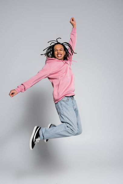full length of positive multiracial man with dreadlocks jumping with outstretched hands on grey  - Foto, Bild