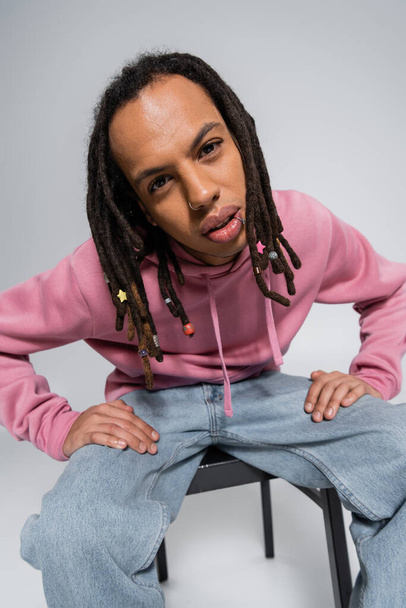 multiracial man with dreadlocks and piercing looking at camera while sitting on chair isolated on grey  - Foto, Imagem