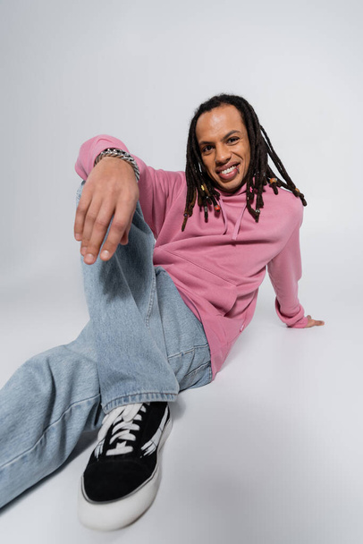 cheerful multiracial man in blue jeans and pink hoodie sitting on gray  - Foto, afbeelding