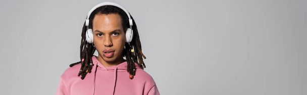 multiracial man with dreadlocks listening music in wireless headphones isolated on grey, banner  - Photo, image