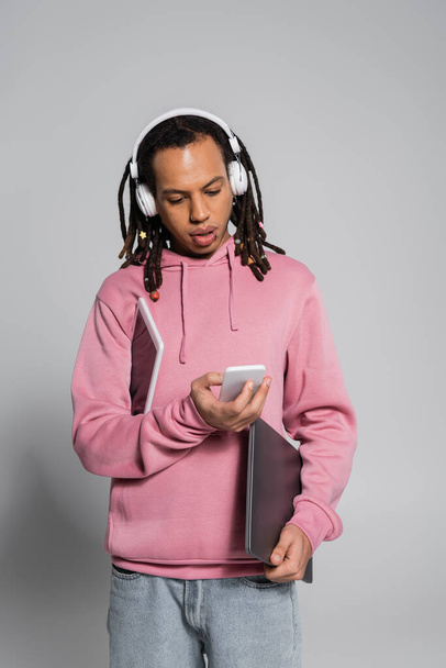multiracial man in wireless headphones using smartphone and holding devices on grey  - Fotoğraf, Görsel