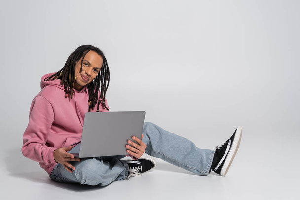 homme multiracial heureux en sweat à capuche rose et jeans à l'aide d'un ordinateur portable tout en étant assis sur fond gris  - Photo, image