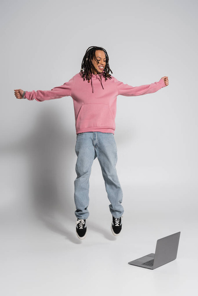 volledige lengte van gelukkig multiraciale man in roze hoodie en jeans springen in de buurt laptop op grijs  - Foto, afbeelding